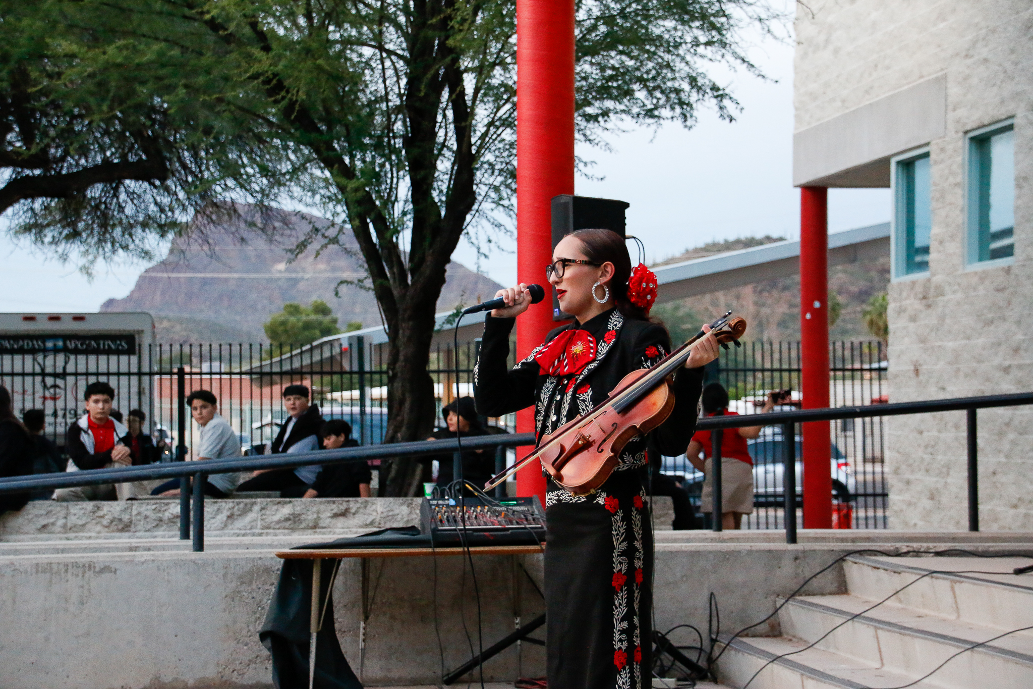 A girl in traditional mariachi costume holds the mic and sings, while holding her violin