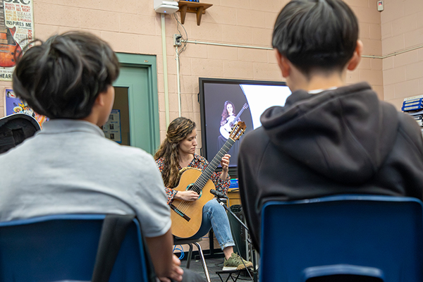 Middle school students listen to a woman give them a guitar lesson